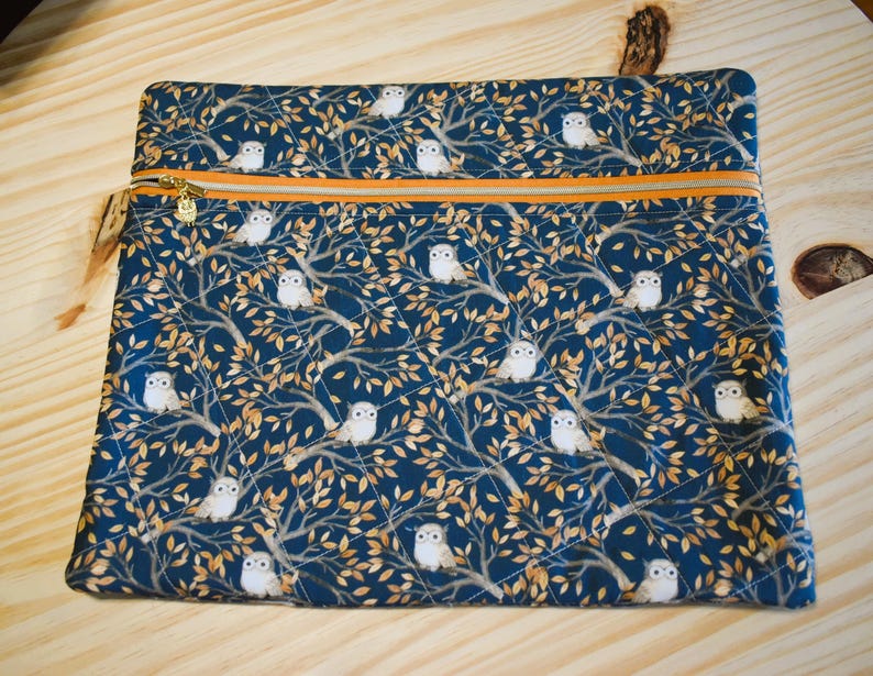 K&ouml;nnte beinhalten: Eine rechteckige, gesteppte Tasche mit marineblauem Hintergrund und einem Muster aus wei&szlig;en Eulen, die in goldbraunen B&auml;umen sitzen. Die Tasche hat einen goldenen Rei&szlig;verschluss mit orangefarbener Paspel und eine kleine, beige Quaste.