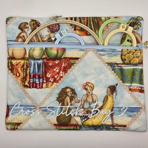 May include: A rectangular quilted fabric bag with a light blue zipper closure, featuring a beach-themed print of people and fruit. Inside the bag are three circular embroidery hoops in light wood, light blue, and light green. Text reads Cross Stitch Bag 2.