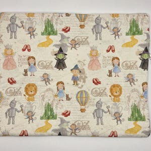 Puede incluir: Una bolsa de tela cuadrada con un patr&oacute;n repetitivo de personajes del Mago de Oz, incluyendo a Dorothy, el Hombre de Hojalata, el Le&oacute;n, el Espantap&aacute;jaros, la Bruja, monos voladores, zapatos rojos, globos aerost&aacute;ticos y la Ciudad Esmeralda.