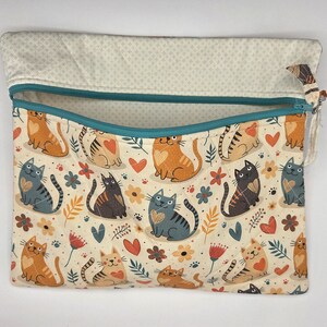 May include: A rectangular fabric pouch with a teal zipper closure. The exterior features a repeating pattern of orange, gray, and black cats interspersed with small red hearts, flowers, and leaves on a cream background. The interior lining is beige.