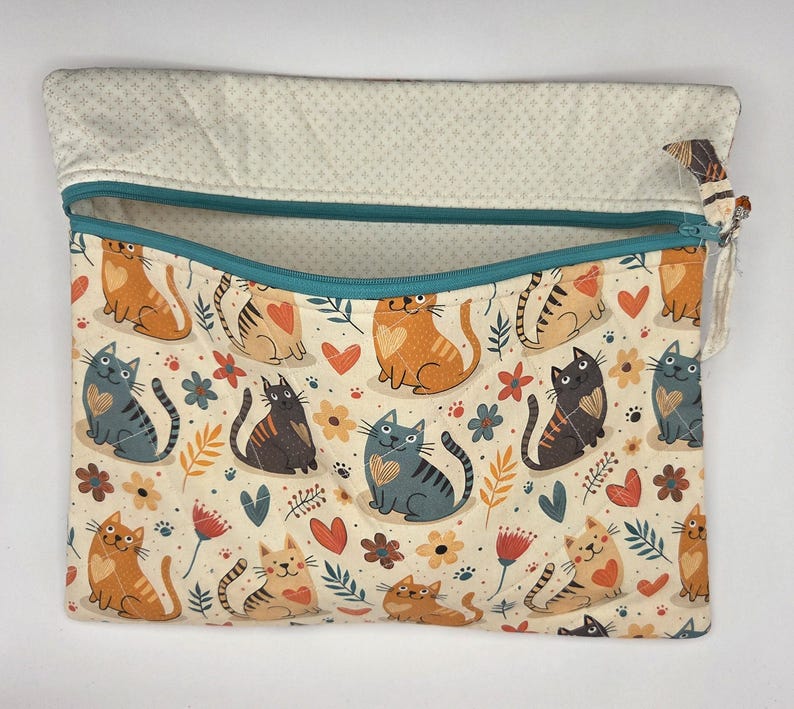 May include: A rectangular fabric pouch with a teal zipper closure. The exterior features a repeating pattern of orange, gray, and black cats interspersed with small red hearts, flowers, and leaves on a cream background. The interior lining is beige.