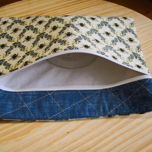 May include: Open zippered pouch with a bee and floral pattern on a cream background. The bottom section is a quilted blue fabric. The interior is a light gray fabric. The pouch is open, revealing the inside.