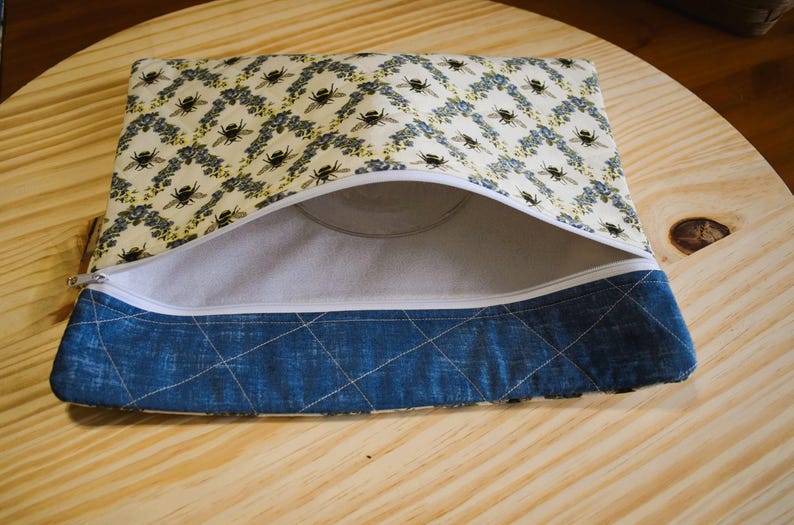May include: Open zippered pouch with a bee and floral pattern on a cream background. The bottom section is a quilted blue fabric. The interior is a light gray fabric. The pouch is open, revealing the inside.