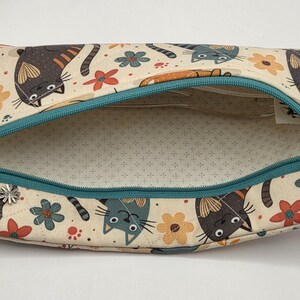 May include: A rectangular fabric zipper pouch featuring a cream-colored background with a repeating pattern of stylized cats in shades of gray, brown, and orange, interspersed with small red and yellow flowers and a teal zipper with a flower charm.