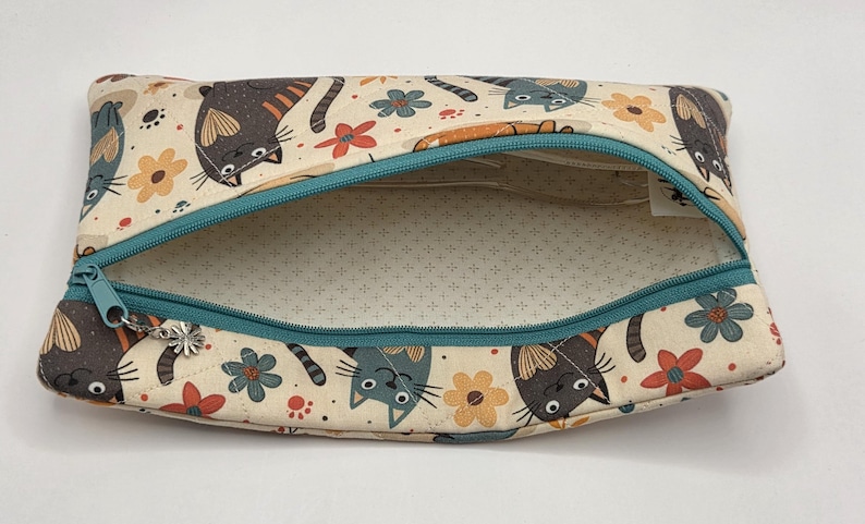 May include: A rectangular fabric zipper pouch featuring a cream-colored background with a repeating pattern of stylized cats in shades of gray, brown, and orange, interspersed with small red and yellow flowers and a teal zipper with a flower charm.