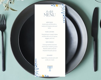 May include: A table setting with a black plate, fork, knife, and spoon. A menu card with the text "Hanukkah MENU" and a list of dishes is centered on the plate. Floral decorations and greenery surround the setting.