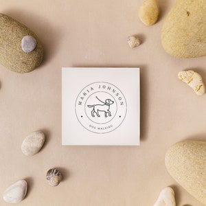 May include: A white card with a black and white logo for a dog walking business. The logo features a dog on a leash and the text "Maria Johnson Dog Walking". The card is surrounded by small stones.