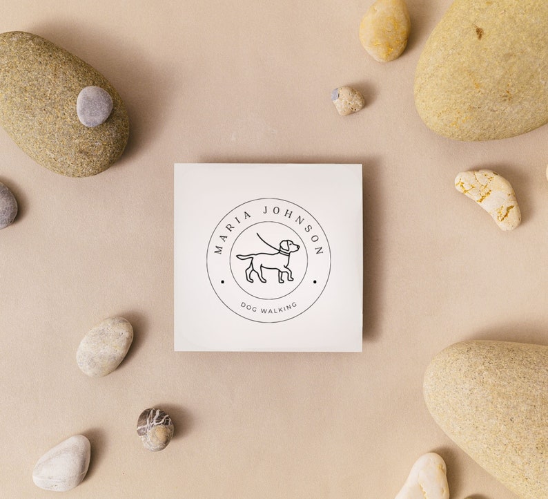 May include: A white card with a black and white logo for a dog walking business. The logo features a dog on a leash and the text "Maria Johnson Dog Walking". The card is surrounded by small stones.
