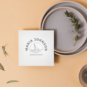 Puede incluir: Una tarjeta blanca con un logotipo en blanco y negro que dice "MARIA JOHNSON Sailing Instructor" con un velero y olas en el logotipo. La tarjeta est&aacute; sobre una superficie marr&oacute;n claro con un plato gris y una planta verde.