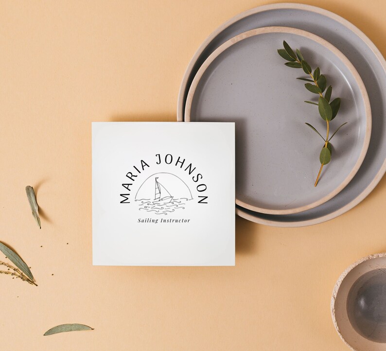 Puede incluir: Una tarjeta blanca con un logotipo en blanco y negro que dice "MARIA JOHNSON Sailing Instructor" con un velero y olas en el logotipo. La tarjeta est&aacute; sobre una superficie marr&oacute;n claro con un plato gris y una planta verde.