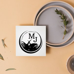 Może przedstawiać: Biała karta z czarnym logo w kształcie koła, na kt&oacute;rym widnieje napis "Lawn Care Service" z inicjałami "MJ" w środku. Logo jest otoczone stylizowanym obrazem trawy.