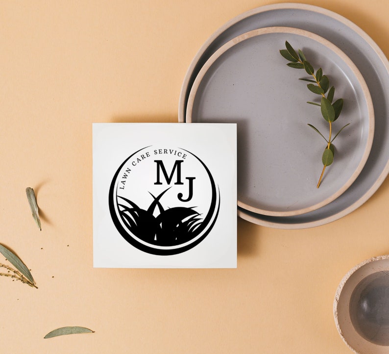 Może przedstawiać: Biała karta z czarnym logo w kształcie koła, na kt&oacute;rym widnieje napis "Lawn Care Service" z inicjałami "MJ" w środku. Logo jest otoczone stylizowanym obrazem trawy.