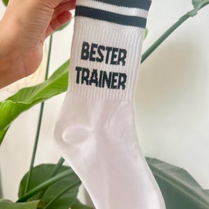 Op de afbeelding: Witte sportsok met twee zwarte strepen aan de bovenkant. De woorden "BESTER TRAINER" zijn in zwarte blokletters op de voorkant van de sok gedrukt. De sok wordt door een hand omhoog gehouden.