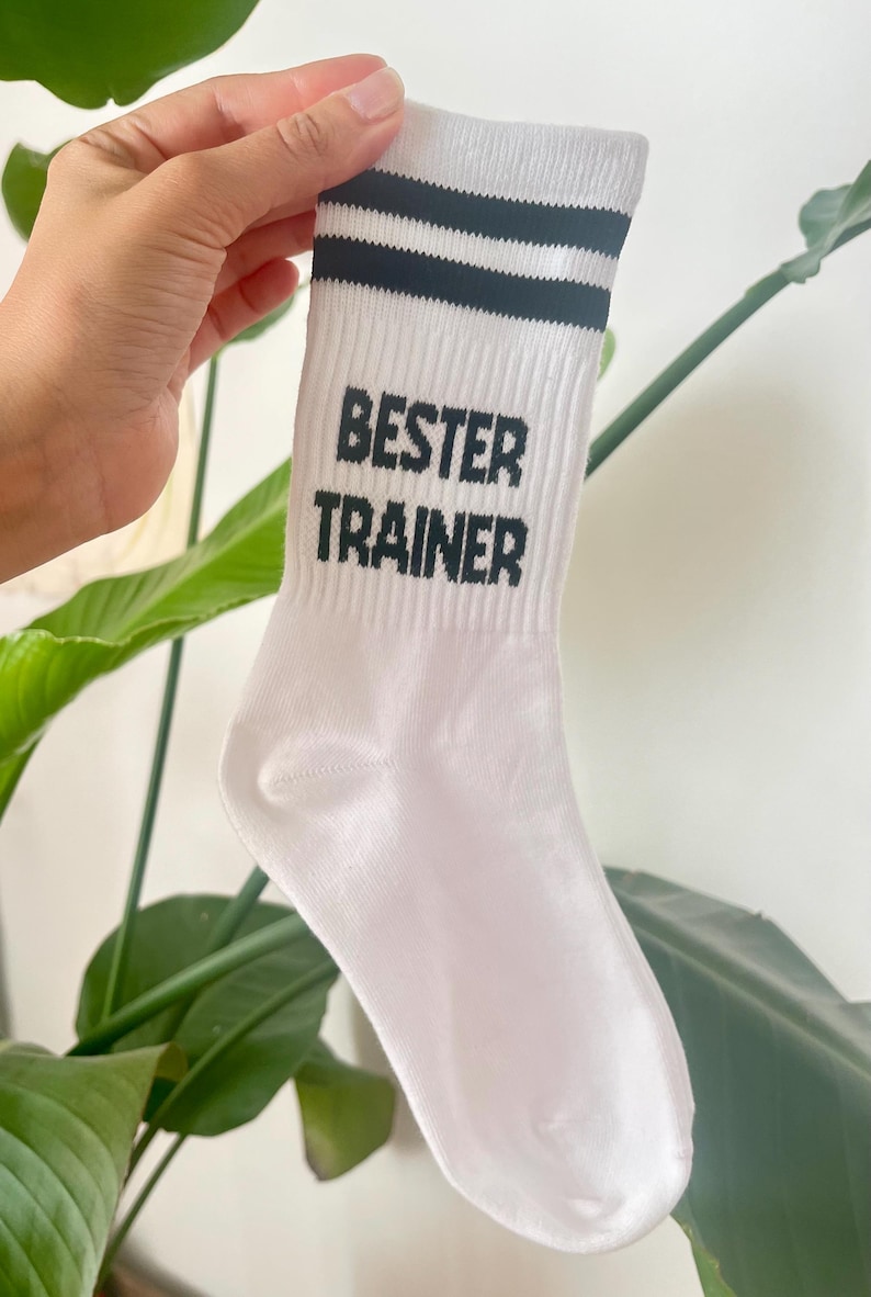 Op de afbeelding: Witte sportsok met twee zwarte strepen aan de bovenkant. De woorden "BESTER TRAINER" zijn in zwarte blokletters op de voorkant van de sok gedrukt. De sok wordt door een hand omhoog gehouden.