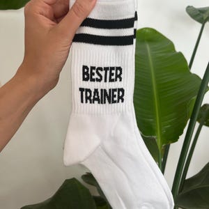 Op de afbeelding: Witte sportsok met twee zwarte strepen aan de bovenkant en de zwarte tekst "BESTER TRAINER". De sok wordt omhoog gehouden tegen een achtergrond van groene bladeren.