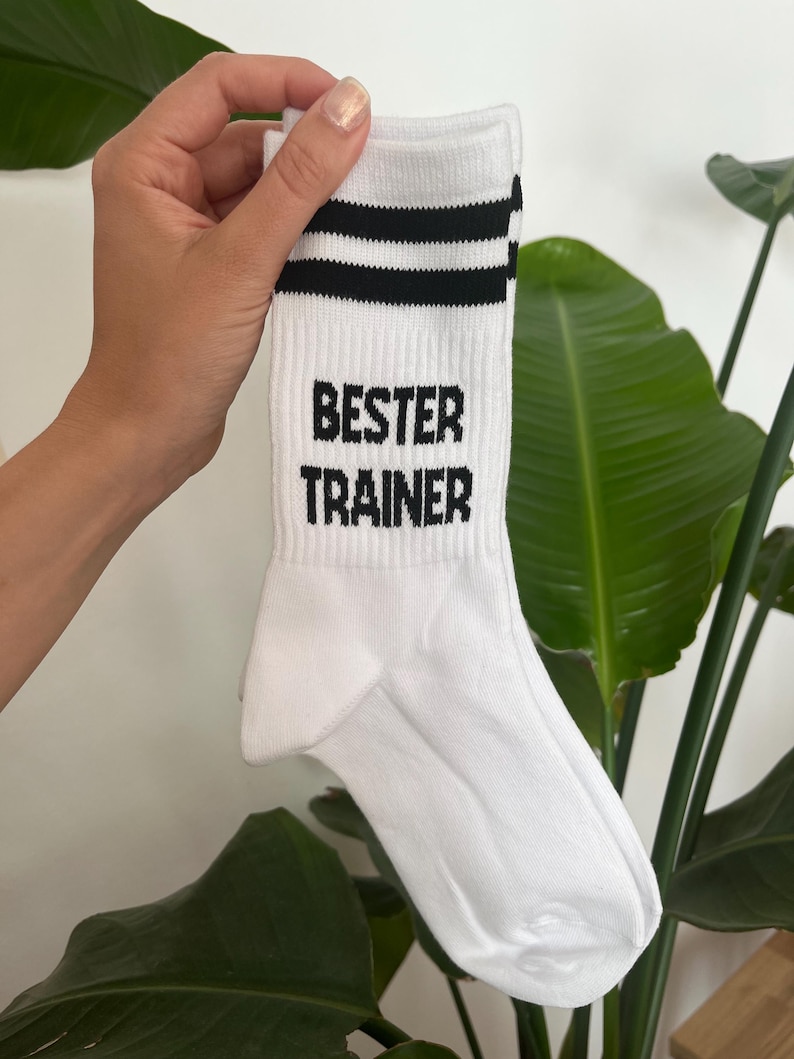 Op de afbeelding: Witte sportsok met twee zwarte strepen aan de bovenkant en de zwarte tekst "BESTER TRAINER". De sok wordt omhoog gehouden tegen een achtergrond van groene bladeren.