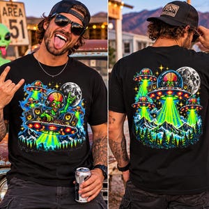 May include: A black t-shirt featuring a vibrant graphic print of a green alien DJ wearing headphones, spinning records on a UFO above a forest and mountain landscape. Multiple smaller UFOs hover in the night sky alongside a full moon and stars.