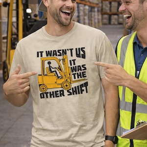 May include: Beige t-shirt with the text "IT WASN'T US IT WAS THE OTHER SHIFT" and a yellow forklift graphic. Two people are pointing at the shirt, smiling. The background shows a warehouse setting with shelves.