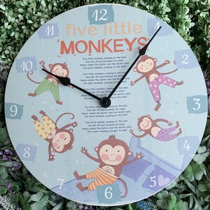 Peut inclure: Une horloge ronde avec un cadran bleu clair représentant cinq singes et le texte "five little monkeys". L'horloge a des aiguilles noires et des chiffres sur le bord.