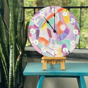 Może przedstawiać: Okrągły zegar z abstrakcyjnym, kolorowym wzorem. Tarcza zegara przedstawia fioletowe, pomarańczowe i niebieskie pociągnięcia pędzla. Białe cyfry oznaczają godziny. Zegar jest prezentowany na małym drewnianym sztaludze. Średnica wynosi około 30 cm.