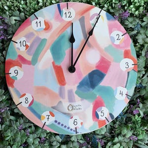 Może przedstawiać: Okrągły zegar ścienny z abstrakcyjnym wzorem w odcieniach różu, niebieskiego, pomarańczowego i zielonego. Tarcza zegara ma czarne wskazówki i białe cyfry na kolorowych kółkach. Zegar ma napis "Clocky Kicks".