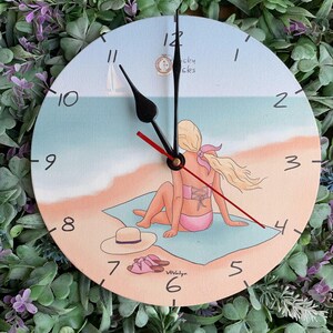 Op de afbeelding: Ronde wandklok met een strandscène. De wijzerplaat toont een vrouw in een roze bikini op een blauw handdoek, met een hoed en sandalen. De klok heeft zwarte wijzers en een rode secondewijzer. De achtergrond toont een zeilboot en de tekst "Lucky Clocks".