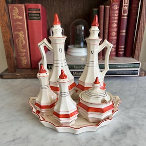 May include: A vintage white ceramic condiment set with red accents, including a tray, two tall pitchers, two smaller bottles, and a lidded sugar bowl. The set features geometric shapes and a red trim. The pitchers have handles and the set is displayed on a marble surface.