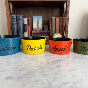 May include: Four colorful snack bowls: blue for "Potato Chips", yellow for "Pretzels", orange for "Snacks", and green for "Nuts". Each bowl has a black interior and a curved shape. The bowls are arranged on a marble surface.