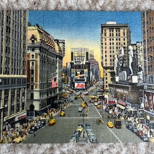 May include: A vintage postcard depicting a busy street scene in New York City. The image shows tall buildings with neon signs, including a large sign for "Camel" cigarettes. Cars and pedestrians are visible on the street.