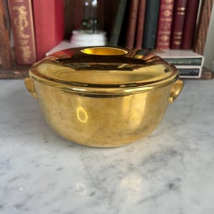 May include: A gold-colored, lidded bowl with two small handles on the sides. The bowl has a rounded shape and a shiny, reflective surface. The lid has a small opening in the center. The bowl is sitting on a marble surface.