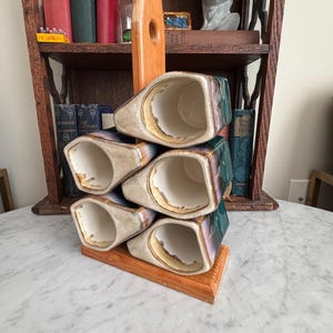 May include: A stack of four ceramic mugs in a wooden holder. The mugs are cream-colored with a gold rim and a green and purple glaze. The wooden holder is a light brown color.