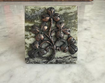 May include: A square green and gray marble paperweight featuring a raised, metallic copper-toned relief of a four-leaf clover plant with multiple leaves and a central stem, centered on the polished stone surface.