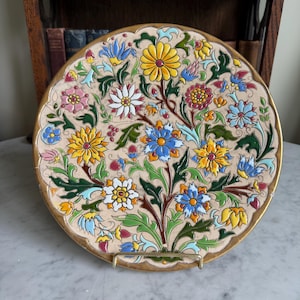 May include: A decorative ceramic plate with a floral design. The plate has a beige background with colourful flowers in yellow, blue, pink, and white. The rim is gold-coloured, and the plate is displayed on a gold-coloured stand.