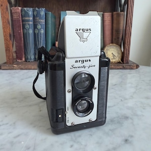 May include: A black and silver Argus Seventy-five box camera featuring a top-mounted viewfinder, a front-facing lens labeled Argus Lumar 75mm Made in U.S.A., and a black carrying strap. The camera body has a textured grip and a shutter setting switch.