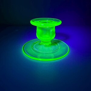 May include: A glowing green glass candlestick holder with a circular base and a flared top. The candlestick holder is illuminated against a dark blue background, showcasing its vibrant color and design. The glass appears to have a slight texture.
