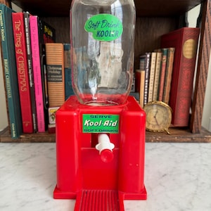 May include: A vintage red Kool-Aid soft drink cooler with a clear glass container. The cooler has a white spigot and a green and white label that reads "Serve Kool-Aid Soft Drinks." The glass container has a green and white label that reads "Soft Drink Kooler."