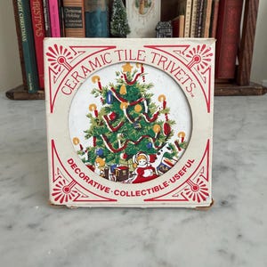 May include: A ceramic tile trivet in a box. The trivet features a Christmas tree design with ornaments and red ribbon. The box is cream-colored with red text that reads "CERAMIC TILE TRIVETS" and "DECORATIVE - COLLECTIBLE - USEFUL."
