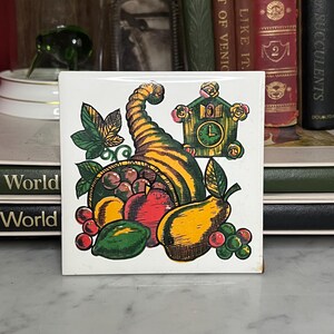 May include: Square ceramic tile with a colorful illustration of a cornucopia overflowing with fruits and a cuckoo clock. The tile features a white background and a vintage aesthetic, perfect for kitchen decor.