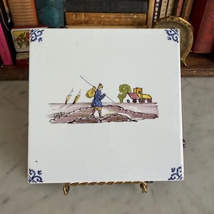 May include: Square white ceramic tile with a hand-painted scene of a person walking with a staff, carrying a bag, and passing by a church. The tile has blue floral corner accents.