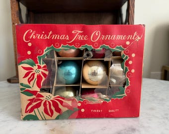 Pode incluir: Uma caixa de cartão retangular contendo vários enfeites de Natal redondos. A caixa vermelha apresenta uma janela recortada, ilustrações de folhas de azevinho verdes e o texto Christmas Tree Ornaments e Finest Quality em letras brancas.