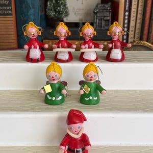 May include: A collection of vintage Christmas ornaments. The ornaments include figures of women in red dresses and aprons, angels in green dresses, and a red-suited elf. The ornaments are painted with details and have gold string hangers.