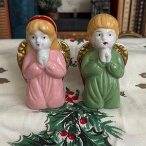 May include: Two ceramic angel figurines, one in pink and the other in green, with gold-colored wings. Both angels have blonde hair, red lips, and are kneeling with hands clasped in prayer. The figurines are on a festive tablecloth.