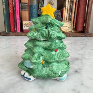 May include: A ceramic Christmas tree-shaped container with a yellow star on top. The green tree has painted details, including colorful lights and a blue and white base. The container is likely used for storage or decoration.