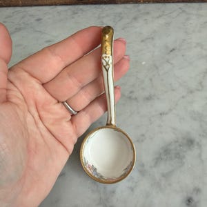 May include: A small, white porcelain spoon with a gold-colored handle and rim. The spoon's bowl features a delicate floral pattern in pink, blue, and yellow. The handle has a decorative design. A vintage-style serving piece.