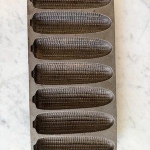 May include: A cast iron cornbread pan with seven corn-shaped molds. The pan is dark gray and has a handle at the top. The molds are detailed to resemble ears of corn, perfect for baking cornbread in the shape of corn.