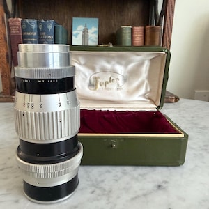 May include: A vintage camera lens with black and silver components, featuring measurement markings. The lens is positioned in front of an open green case with a red velvet interior and a white satin lining. The case has the word "Toplen" printed on it.
