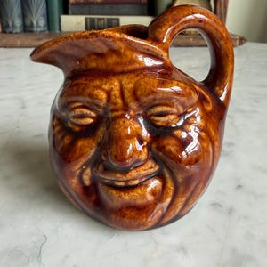 May include: A brown ceramic pitcher with a smiling face design. The pitcher has a handle and a spout. The face has closed eyes and a wide, smiling mouth. The ceramic has a glossy finish.