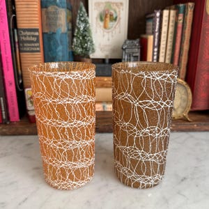 May include: Two vintage amber-colored drinking glasses with a white, web-like design. The glasses are cylindrical and stand upright, showcasing a retro aesthetic. They are placed on a marble surface, with books and decorative items in the background.