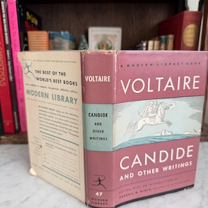 May include: A vintage copy of "Candide and Other Writings" by Voltaire, a Modern Library book. The book has a pink spine with white text and a light blue cover illustration. The book is displayed with other books on a shelf.