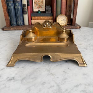May include: A vintage brass inkwell with two ink pots and a central trough. The inkwell has an ornate, Art Nouveau-inspired design with a decorative top. The inkwell is displayed on a marble surface, with books and a clock in the background.